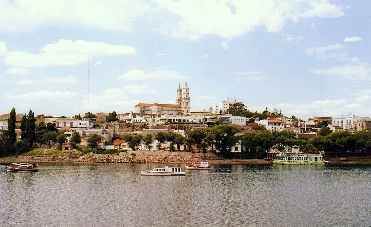 Wide river under a blue sky with small boats, and on the far bank a low town of white houses and trees with a twin-towered church