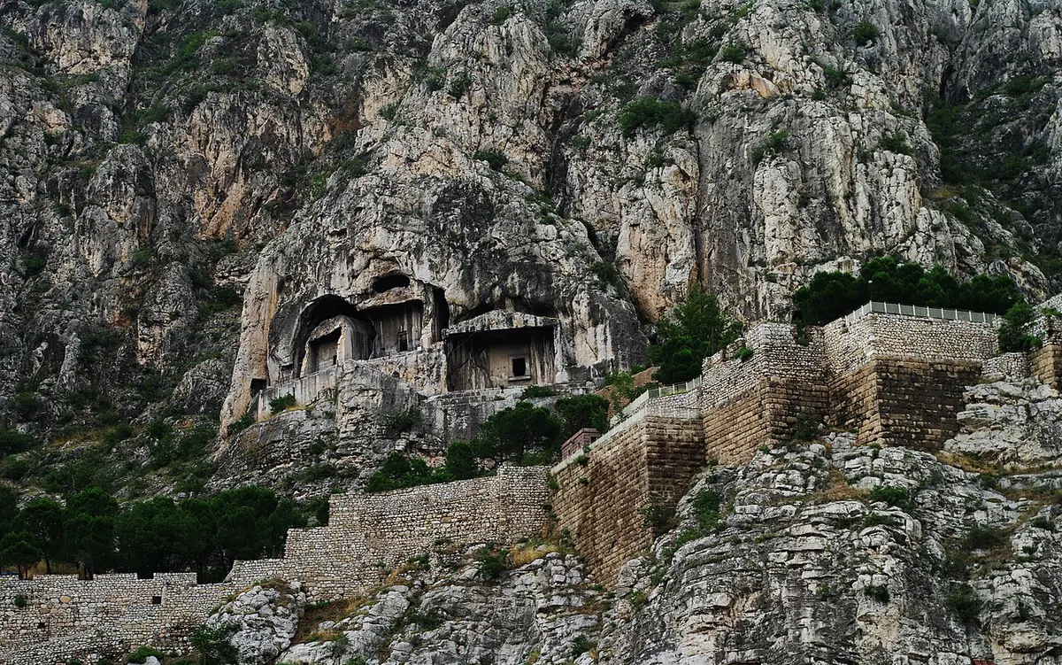 Ancient tombs with columned facades carved into a towering gray cliff above old fortification walls at Amasya, Turkey