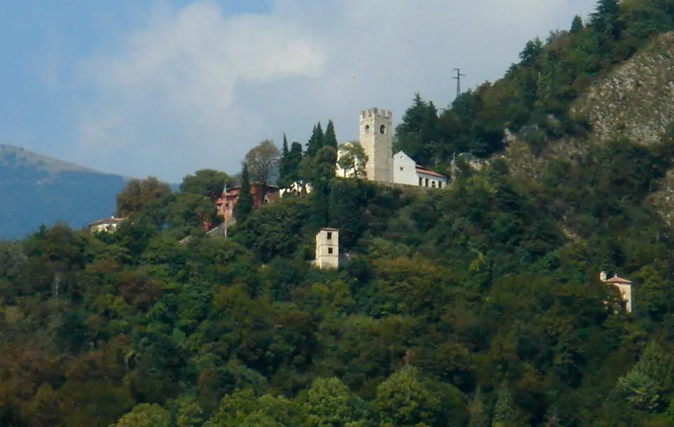 A white church and square stone bell tower among green trees high on a steep wooded hillside, under a blue sky