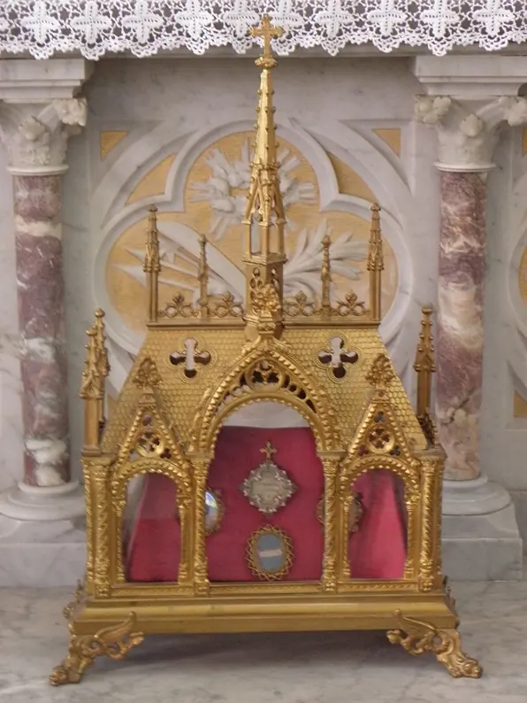 A gilded neo-Gothic reliquary shaped like a small chapel with pinnacles and glass windows, holding relics in oval frames on red silk, on a white marble altar
