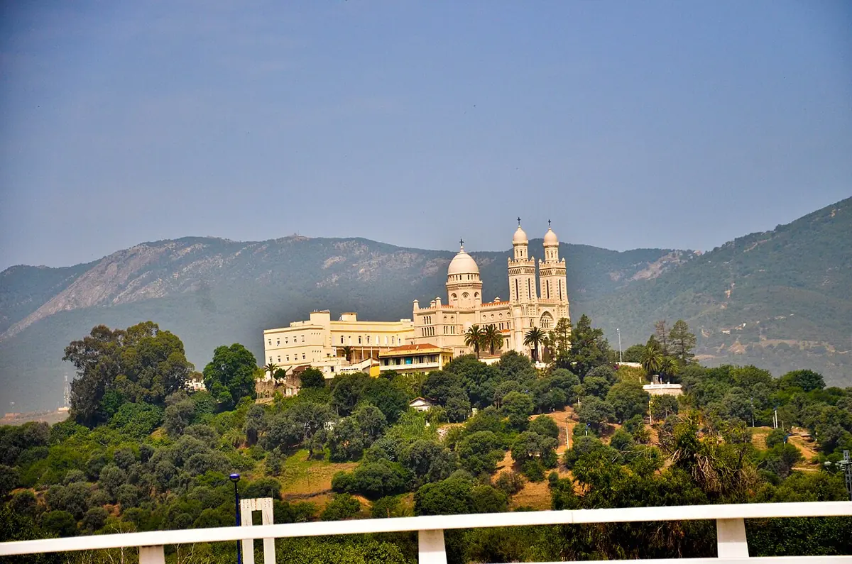 Cream-colored basilica with twin towers and a dome on a wooded hill above Annaba, with mountains behind