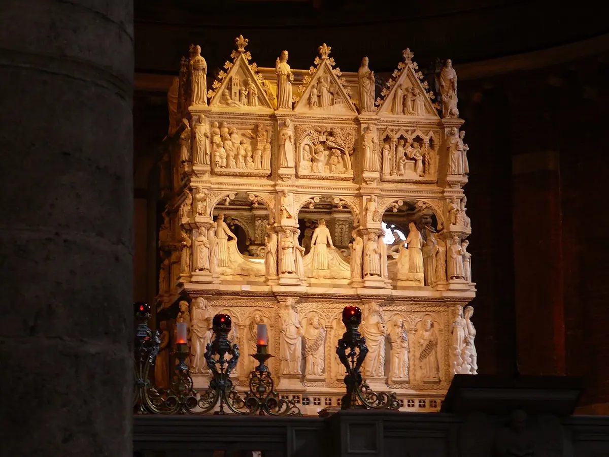 The tall white marble shrine of St. Augustine, carved with dozens of small statues and reliefs, glowing in a dark church sanctuary behind candlesticks