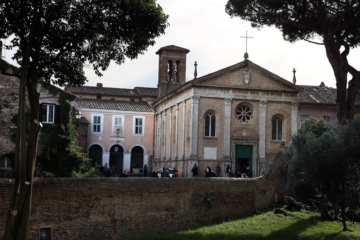 Small Renaissance church of pale stone with a rose window and a brick bell tower, behind a garden wall and umbrella pines