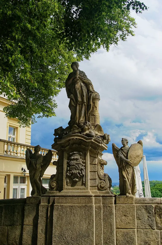 Baroque stone statue of St. Barbara on a tall carved pedestal on a balustrade, with other statues and trees behind it, in the silver-mining town of Kutná Hora