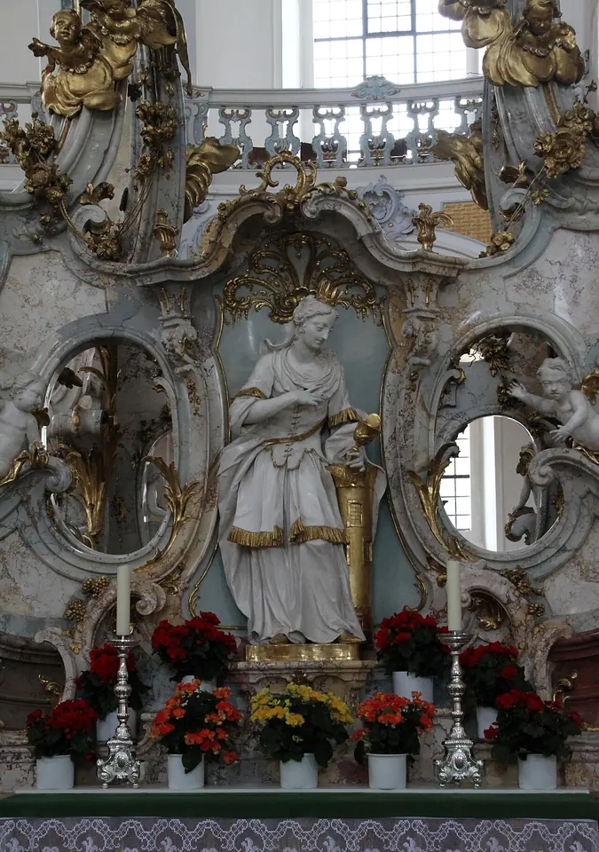 White and gold Rococo statue of St. Barbara holding a chalice beside a tower, set among the arches of the shrine altar at Vierzehnheiligen, with red flowers below