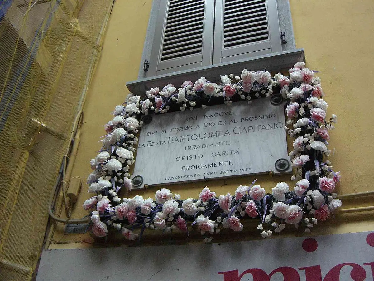 Marble plaque on a yellow house wall, framed by a garland of pink and white flowers, with an inscription honoring Bartolomea Capitanio