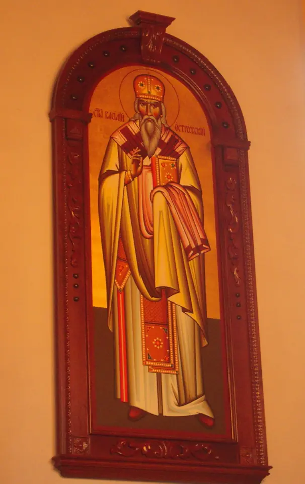 Icon in an arched carved wooden frame of a standing bishop in gold vestments and miter, holding a book, on a warm orange background