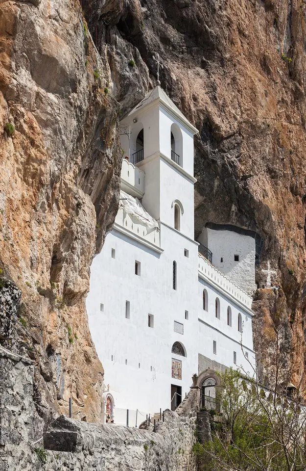 White monastery walls and a bell tower built into a hollow in a towering cliff of gray and orange rock
