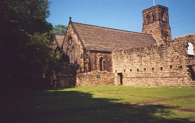 Low ruined stone walls on a lawn in front of a small stone church with a square tower, under a blue sky