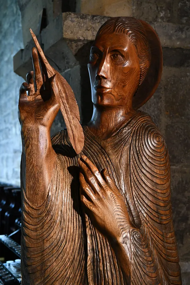 Tall modern wood carving of a monk in a long ribbed robe with one hand raised holding a quill, his face turned upward