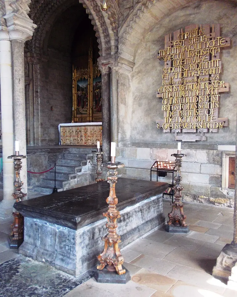 A plain dark stone tomb chest between tall candlesticks under Romanesque arches, with a gold lettered text panel on the wall behind it
