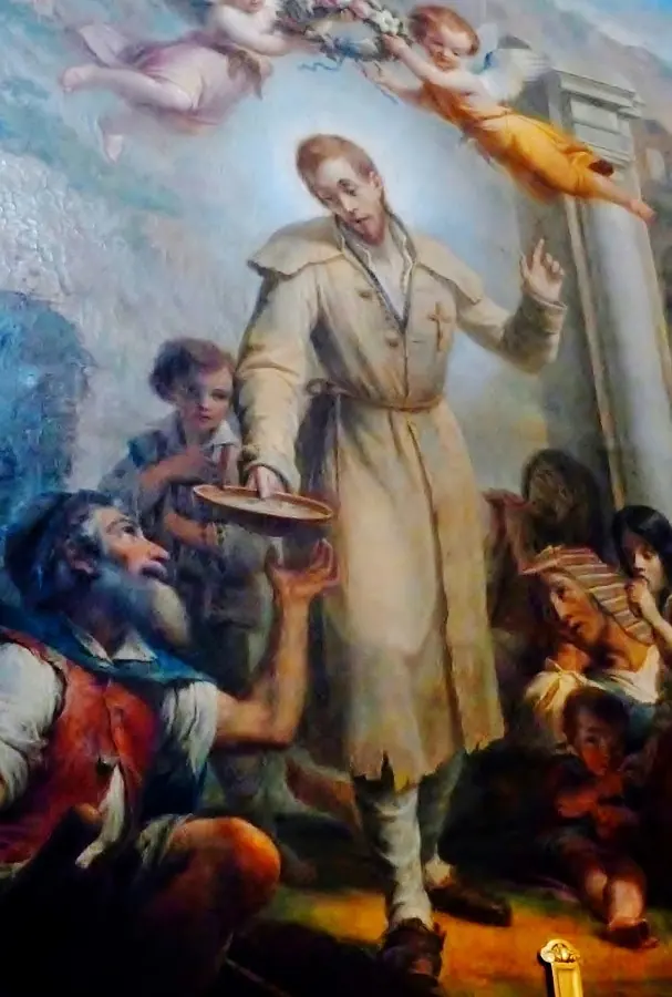 Painting of Labre in a pale coat sharing food from a bowl with poor men and children while cherubs drop flowers from the sky