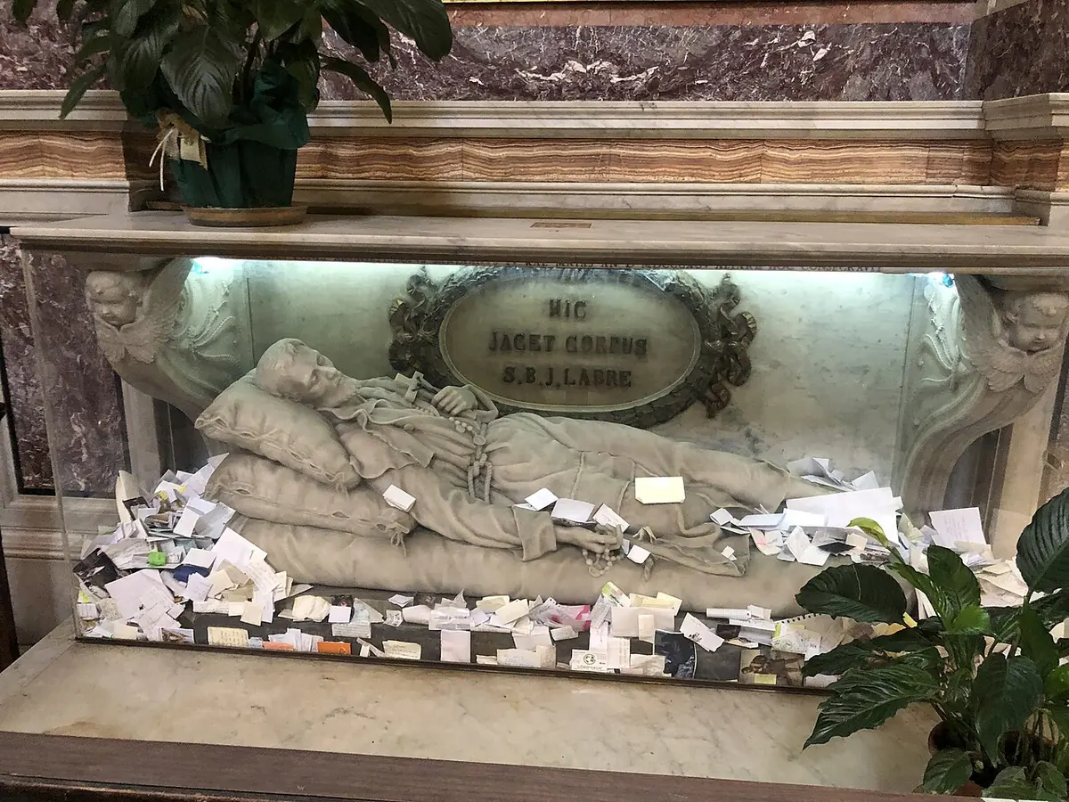 White marble effigy of Labre lying in death under an altar, with dozens of folded paper prayer notes tucked around it