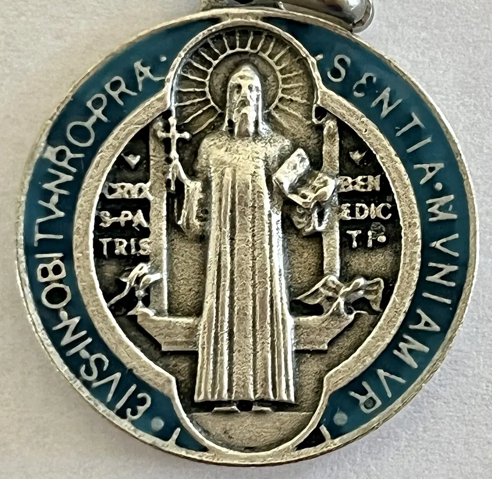 Close-up of the front of a silver and blue enamel St. Benedict medal, the saint standing with a cross and book between the poisoned cup and the raven, with the words Crux S. Patris Benedicti and a Latin inscription around the rim