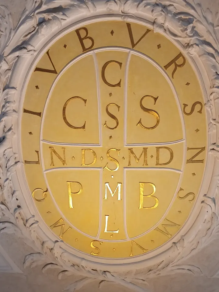 A large oval of the St. Benedict medal's back worked into a ceiling, gold letters CSSML, NDSMD and C S P B on a softly lit cream ground framed by stucco