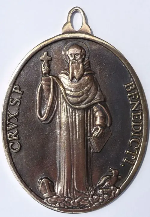 Front of a large oval bronze medal showing St. Benedict holding a small cross and a book, a raven at his feet, with the words Crux S. P. Benedicti around the edge