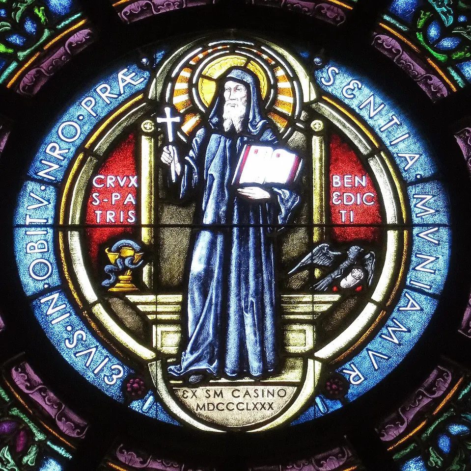 Stained-glass roundel of St. Benedict in a blue habit holding a cross and an open book, with a cup on one side and a raven on the other, and the medal's Latin inscription around the edge