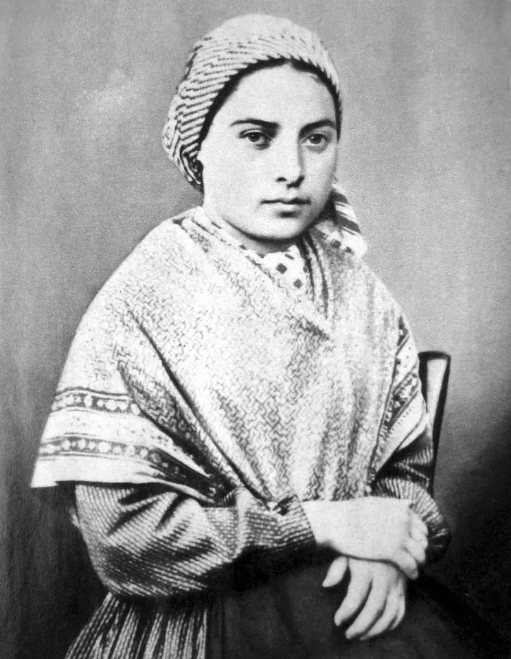 Black-and-white photograph of a teenage girl in a striped headscarf and patterned shawl, seated with folded hands, looking straight at the camera