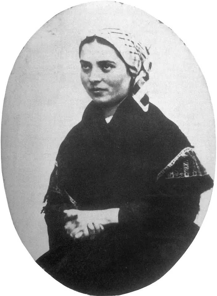 Black-and-white photograph of a young woman in a long white hood falling over a dark shawl and dress, her hands folded, looking to one side