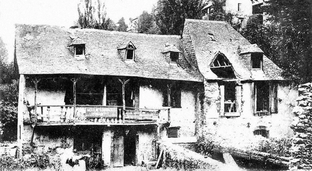 Old postcard photograph of a stone mill house with a wooden balcony, a slate roof with dormer windows and trees around it