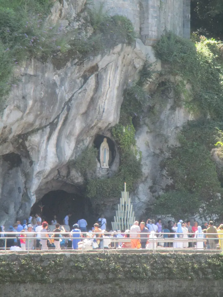 Pilgrims standing at a railing before a gray rock grotto, with a white statue of the Virgin in a niche above and a tall stand of burning candles below