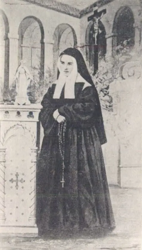 Grainy photograph of a young nun in a black habit and white bonnet standing with a rosary beside a statue of the Virgin in front of an arcade