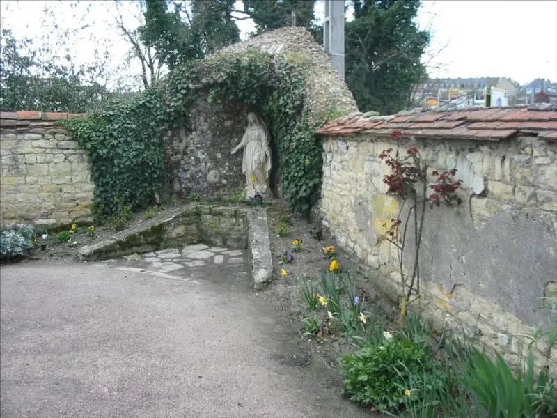A small rock grotto with a white statue of the Virgin set into an ivy-covered stone garden wall, with a gravel path in front
