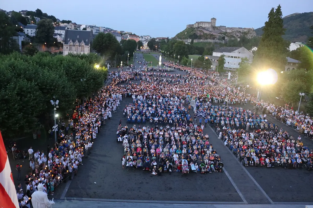 Aerial view at dusk of thousands of pilgrims holding candles in a vast square in front of the Lourdes basilicas