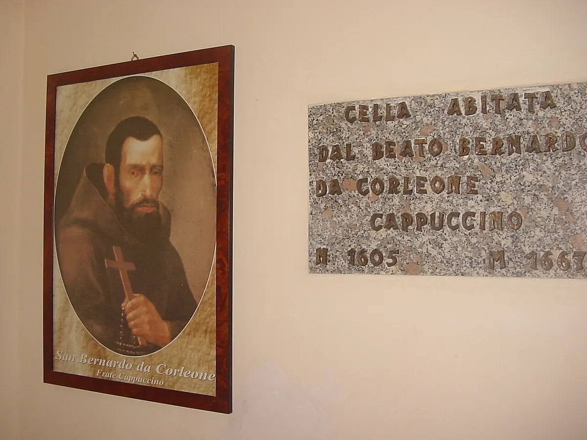 A framed portrait of a bearded Capuchin holding a crucifix hangs on a plain wall beside a stone plaque marking the cell of Blessed Bernard of Corleone