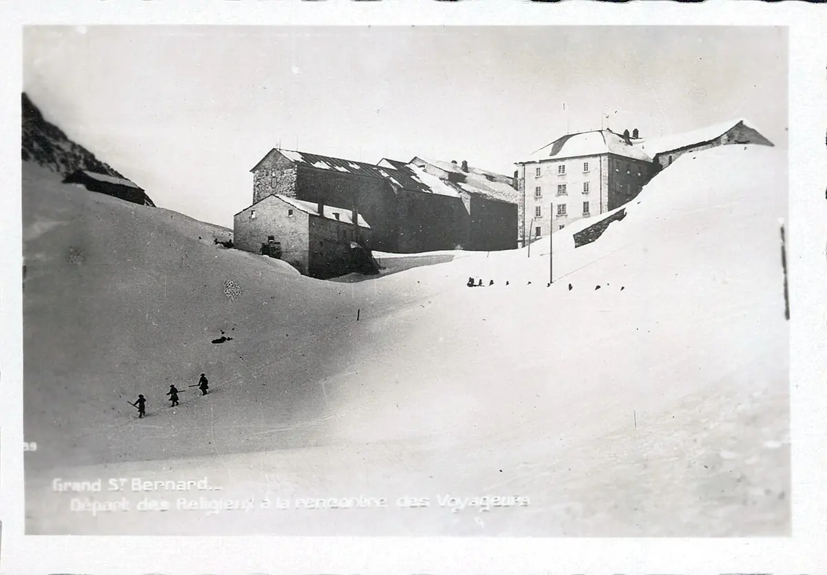 Old black-and-white photograph of the stone hospice buildings half buried in deep snow on the pass, with a few small figures crossing the snow in the foreground