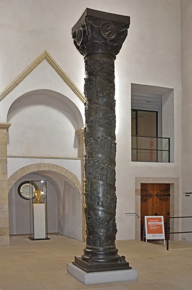 A dark bronze column standing in a white stone church hall, with a band of relief figures spiraling up its shaft to a carved capital