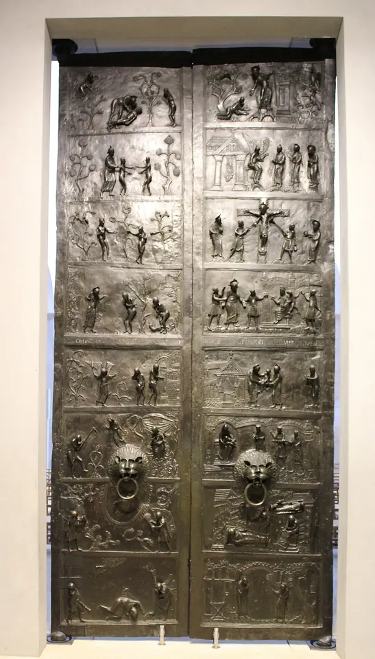 Tall pair of dark bronze doors covered in rows of small relief scenes, with two lion-head door handles in the middle