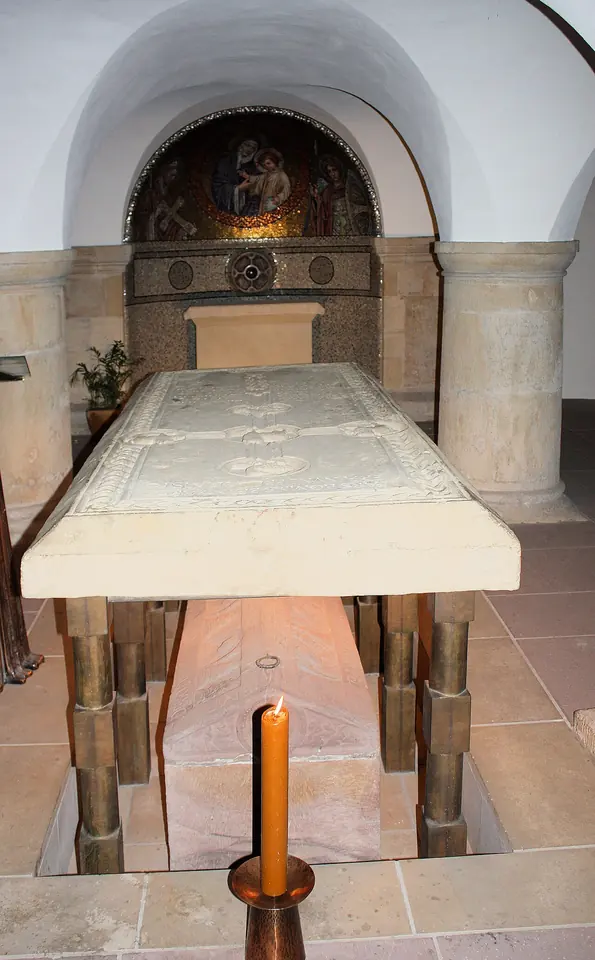 Vaulted stone crypt with a raised stone sarcophagus lid on metal supports, a tall candle in front and a small mosaic of the Virgin in the apse behind