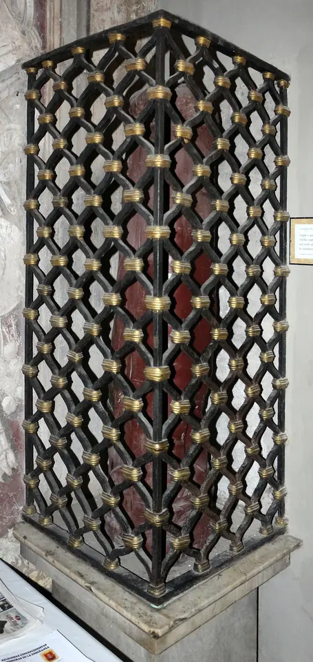 Short red marble column enclosed in a black and gold lattice grille, standing on a stone base inside a church