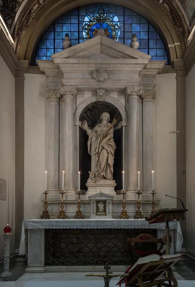 Marble altar niche framed by white columns and a triangular pediment, holding a white statue of St. Bibiana above a lit altar with tall candlesticks