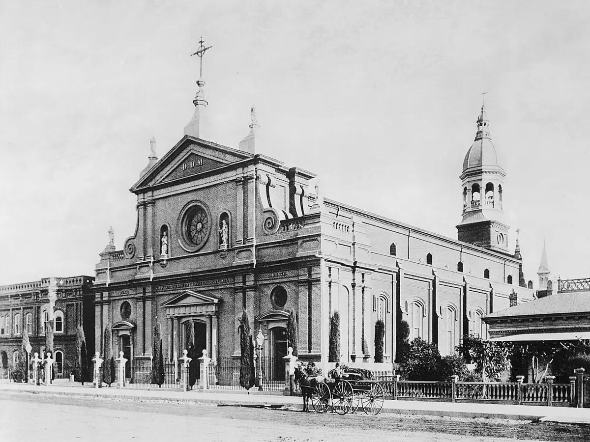 Old photograph of a pale cathedral with a round window and a domed bell tower on a dirt street in early Los Angeles, a horse and buggy in front