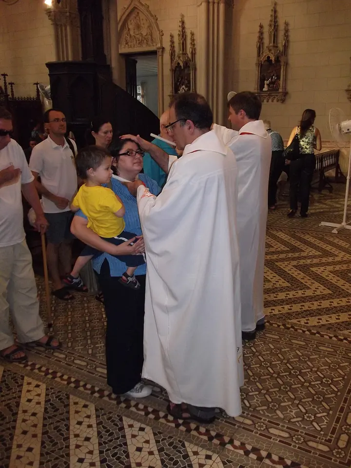 A priest in a white alb touches crossed candles to the throat of a woman holding a small boy, while others wait in line in a church