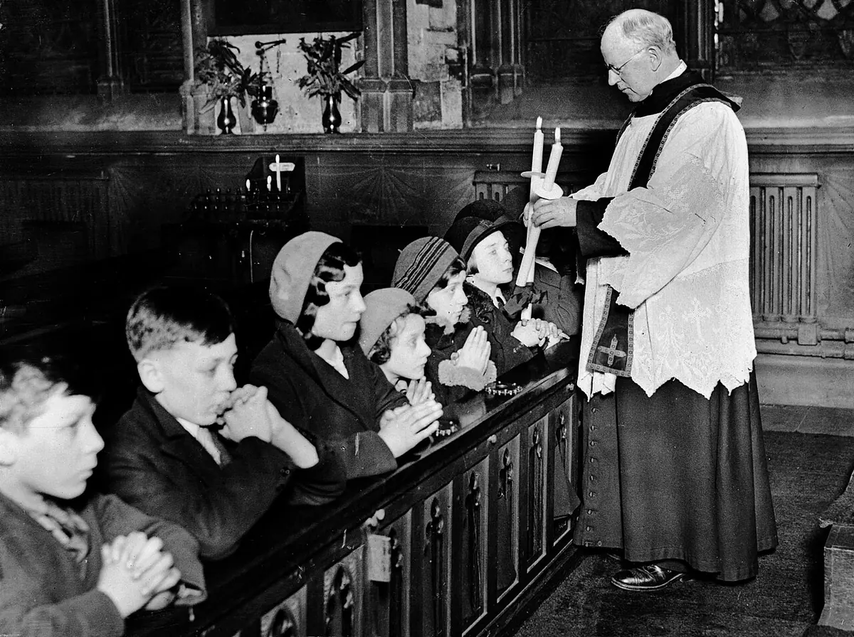 Black-and-white photograph of a priest in a surplice holding two crossed candles at the throat of a boy as a row of children kneels at an altar rail