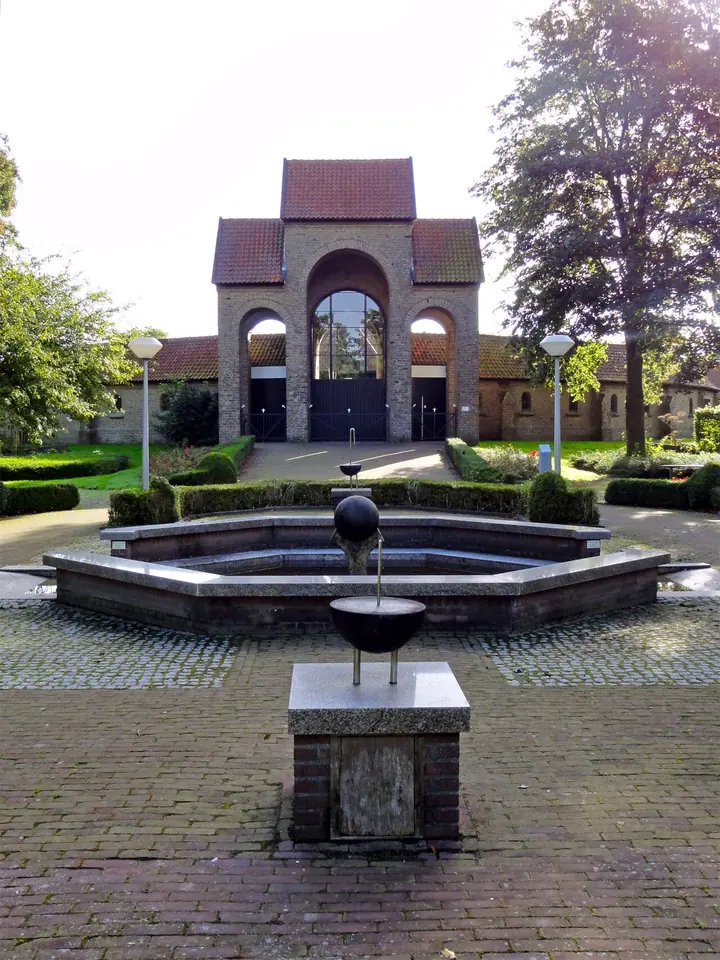 Brick pilgrimage chapel with an open arched entrance behind a round fountain basin in a paved square in Dokkum