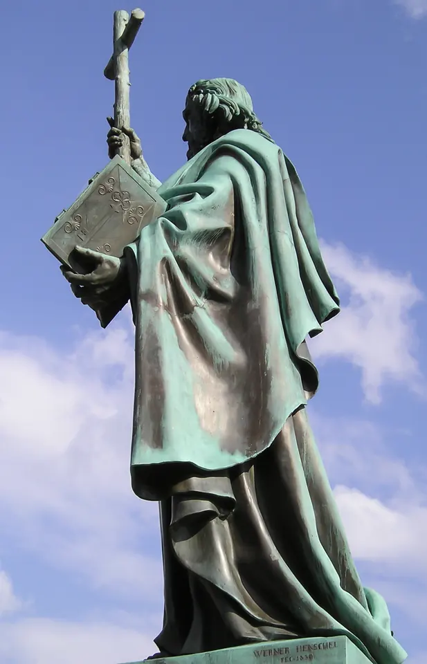Green bronze statue of St. Boniface in a long cloak, holding a cross raised high and a book, against a blue sky
