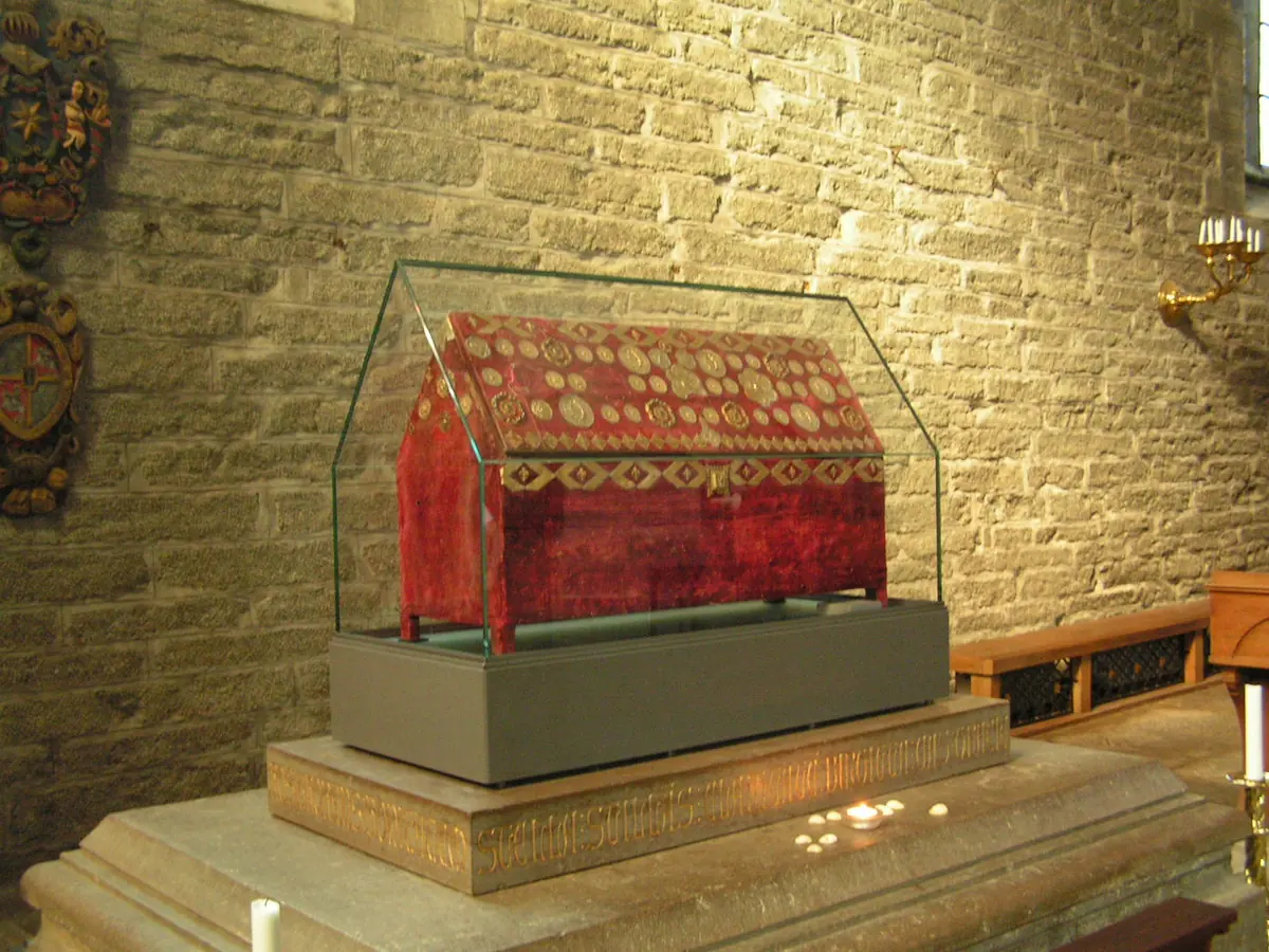 A red velvet-covered reliquary casket decorated with metal ornaments, under a glass case on a stone base against a brick wall
