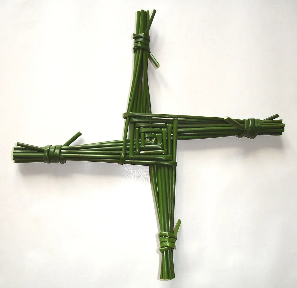 A four-armed St. Brigid's cross woven from green rushes, with a square woven center and the arm ends tied off, against a white wall