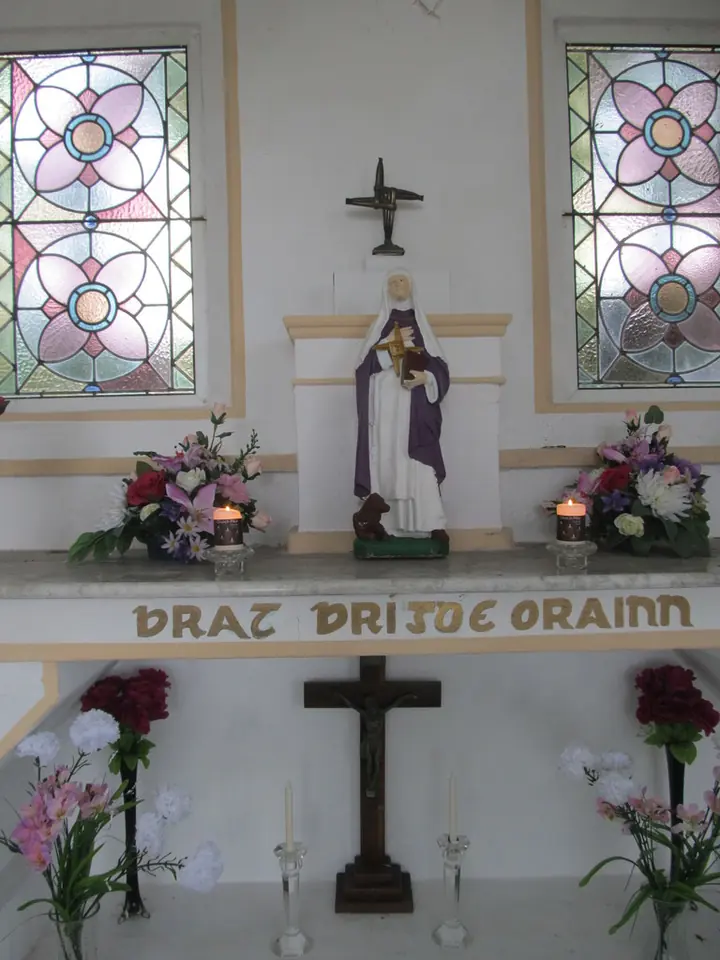A white shrine with a small statue of St. Brigid under a rush cross, flowers and candles on the altar, and the Irish words Brat Bríde orainn painted below