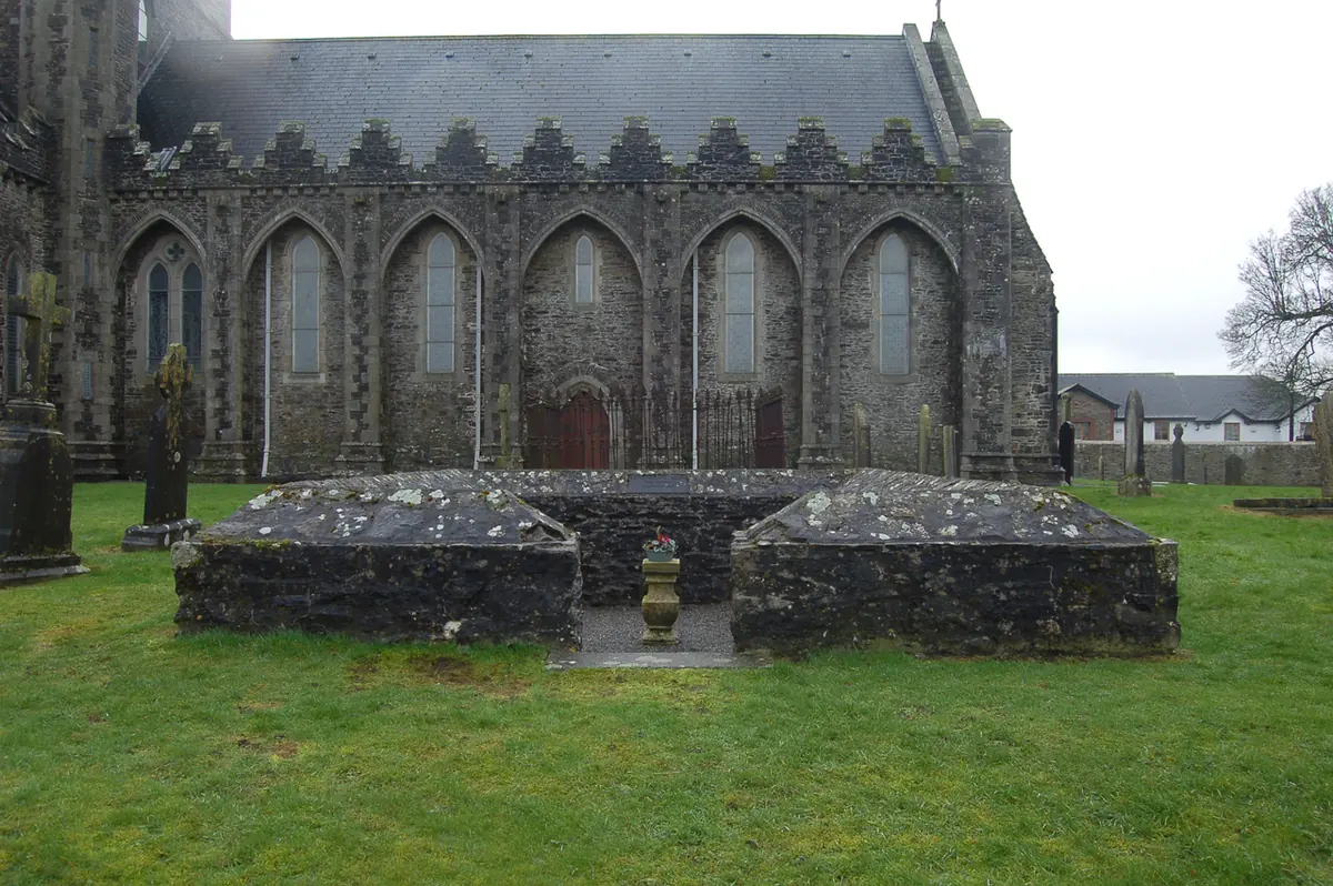 The low rectangular stone foundations called St. Brigid's fire temple on the lawn beside the gray stone wall and tall windows of Kildare Cathedral