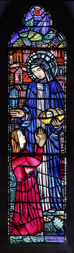 A tall narrow stained-glass window in deep blues and reds showing St. Brigid in a blue mantle with a halo, giving to kneeling figures below, with a church behind her