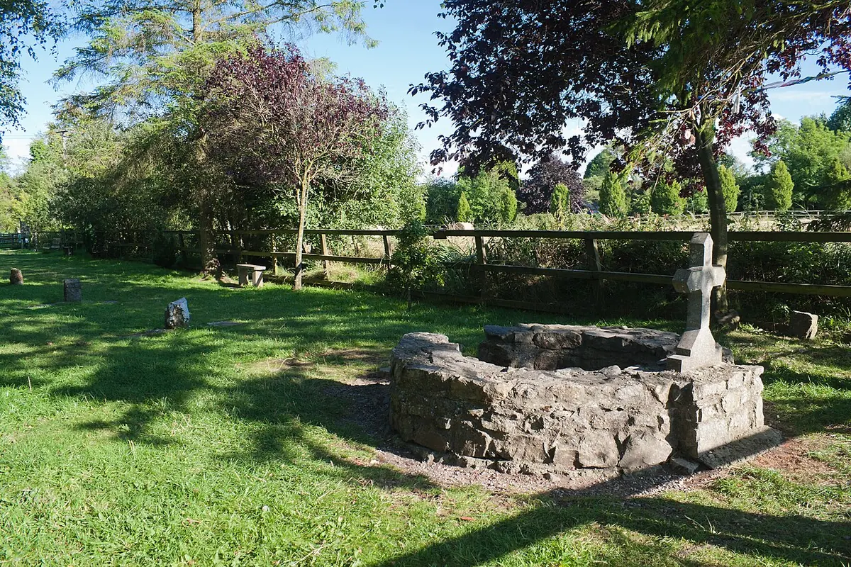 A low circular stone well with a stone cross beside it on a grassy site ringed by trees outside Kildare