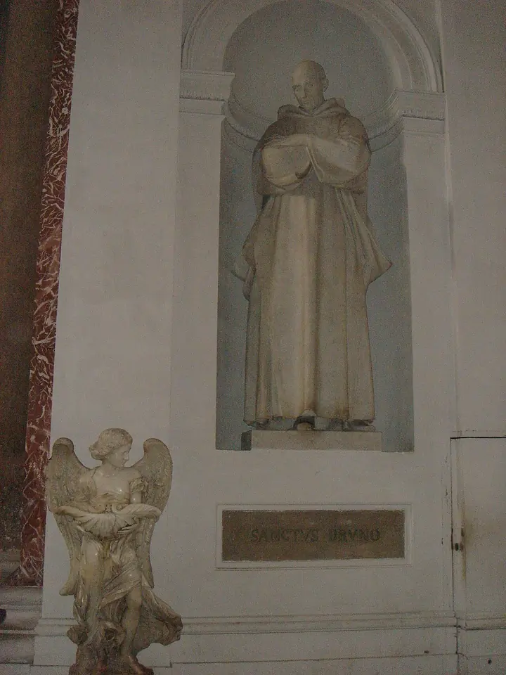 A white marble statue of a monk in a cowl with his arms crossed on his chest, standing in a niche above a marble angel holding a holy-water basin