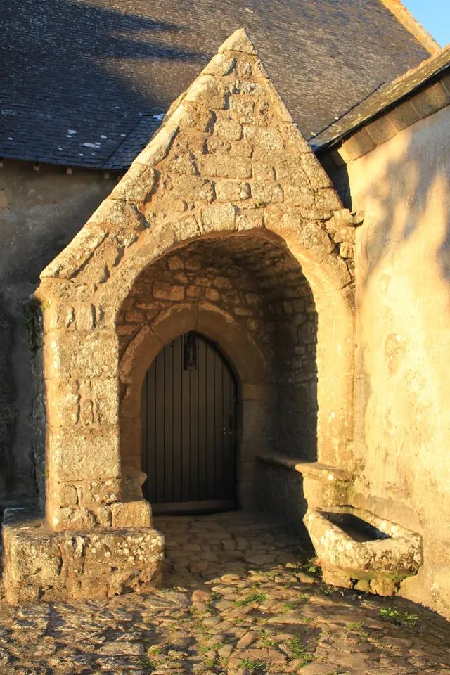 The arched stone porch of a small granite chapel glowing in evening sun, with a stone bench and water basin inside