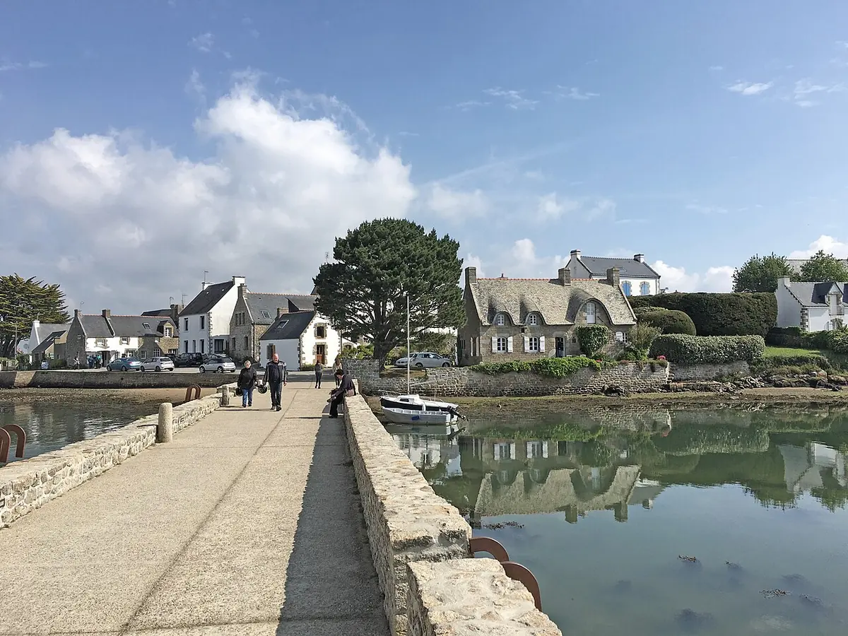 A stone causeway crossing calm water to a tiny island village of stone cottages with a chapel among the trees