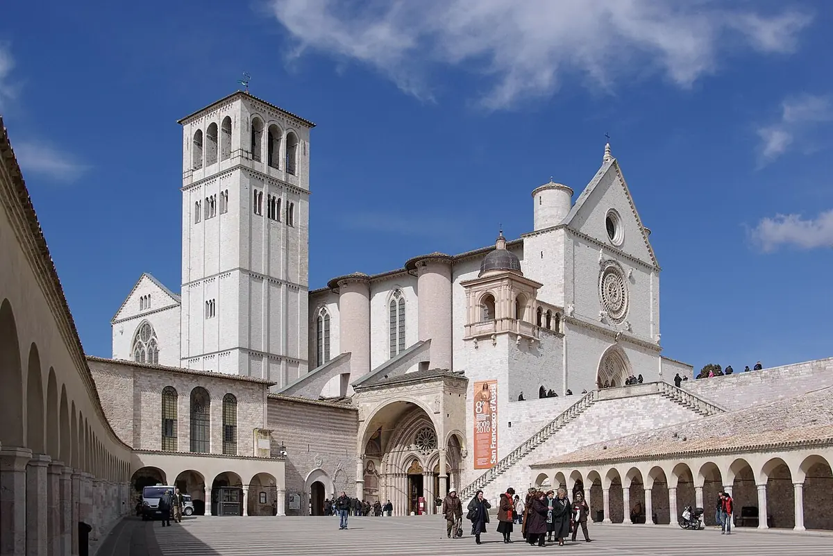 La basílica de San Francisco de Asís, de piedra clara, con su campanario, vista desde la plaza inferior porticada bajo un cielo azul